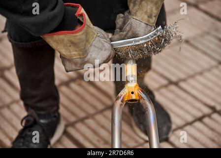 Homme méconnaissable avec des gants de protection à l'aide d'une brosse métallique au-dessus d'une fourche de vélo après avoir passé un chalumeau dessus pour enlever la peinture, dans le cadre de son Banque D'Images