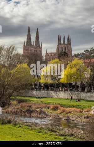 Burgos, Espagne - 25 mars 2023 : vue sur la rivière Arlanzon, les gens assis sur une pelouse, la cathédrale gothique derrière les arbres Banque D'Images