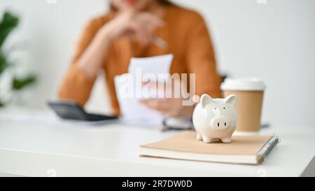 Image rapprochée d'une banque de porc sur une table avec une femme planifiant ses dépenses mensuelles. dépenses, épargne, budget, revenu, prêt immobilier. défoqué i Banque D'Images