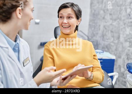 Portrait d'une femme mûre souriante en clinique dentaire parlant à une femme dentiste et tenant une tablette Banque D'Images