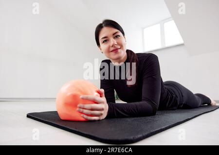 Une jeune fille est engagée à Pilates dans un studio lumineux. Souriant sportswoman dans un costume noir se trouve sur un tapis de yoga et tient une boule orange dans ses mains. H Banque D'Images