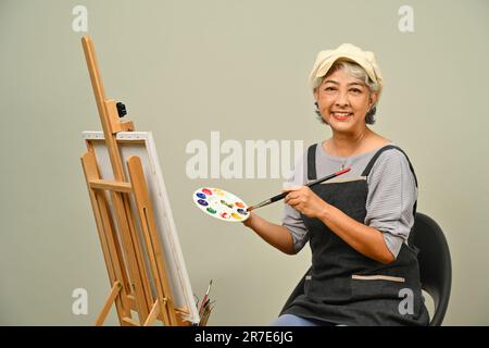 Portrait d'une belle femme âgée de 60s ans portant un tablier tenant la brosse et la palette sur fond gris Banque D'Images