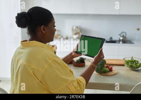 Vue arrière d'une femme afro-américaine regardant la cuisine en ligne sur un Tablet pc assis dans la cuisine Banque D'Images