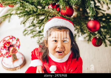 Portrait de Noël d'une adorable petite fille asiatique de 3 ans portant une robe et un chapeau de père Noël rouges, assise sur le sol, vue du dessus Banque D'Images