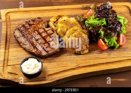 Entrecôte grillée Black Angus servie avec des frites, une salade verte et de la sauce sur fond de bois. Banque D'Images
