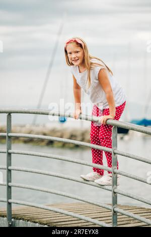 Bonne fille de 9 ans s'amusant dehors, jouant au bord du lac pendant une belle soirée chaude et ensoleillée, portant un t-shirt blanc et des chaussures, un pantalon à pois rouges et h Banque D'Images