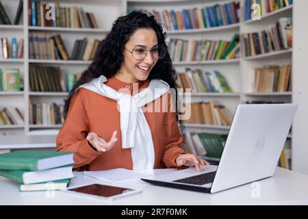 Jeune femme hispanique parlant sur appel vidéo étudiant à l'intérieur de la bibliothèque universitaire, apprentissage en ligne. Banque D'Images