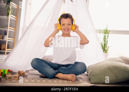 photo de garçon positif s'asseoir camp fait maison dans la salle préscolaire écouter des chansons de dessin animé avec un casque Banque D'Images