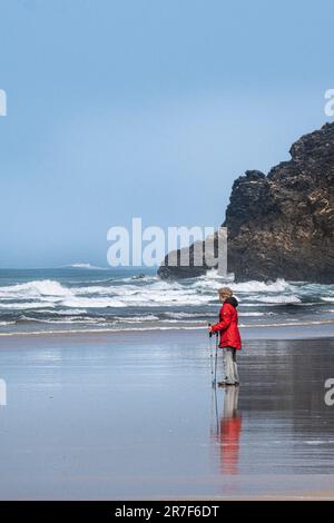 Une femelle mature portant un manteau rouge vif et utilisant des bâtons de marche bâtons de randonnée debout sur le rivage à la plage de Mawgan Porth dans les Cornouailles au Royaume-Uni. Banque D'Images