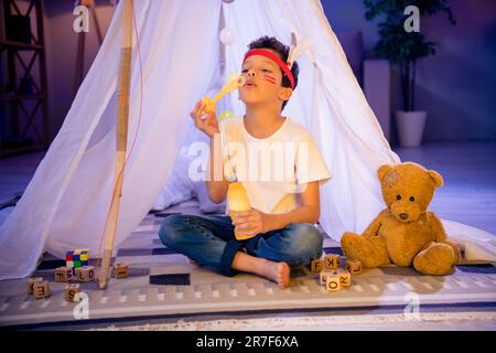 Photo d'un adorable petit garçon dans un costume américain natif. Fête d'anniversaire, un sleepover joue des bulles de savon dans un camp fait maison dans la salle de jeux Banque D'Images