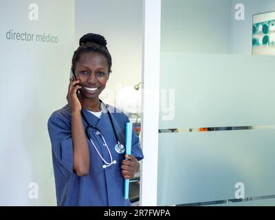 Jeune médecin debout avec son stéthoscope dans son bureau tout en parlant au téléphone Banque D'Images