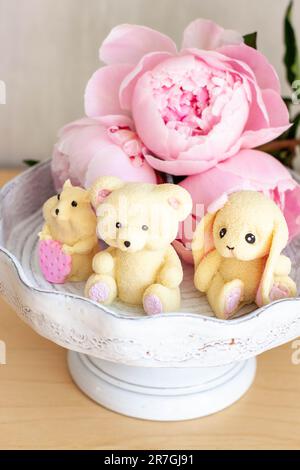 Adorable ours en peluche comestible, lapin et jouets de hamster faits de chocolat blanc avec des pivoines fraîches et des bougies sur les fonds. Cadeau pour bébé Banque D'Images