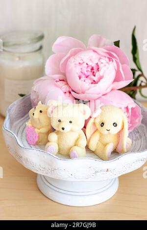 Adorable ours en peluche comestible, lapin et jouets de hamster faits de chocolat blanc avec des pivoines fraîches et des bougies sur les fonds. Cadeau pour bébé Banque D'Images