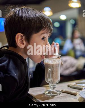 Faire dorer le petit garçon dans un bar en buvant du lait au chocolat chaud avec une cuillère. Arrière-plan hors foyer Banque D'Images