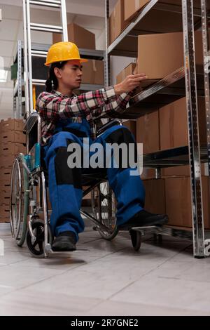 Jeune ouvrier d'entrepôt asiatique utilisant un fauteuil roulant tout en faisant l'inventaire dans la salle de stockage. Le gestionnaire de la logistique qui vérifie les colis de marchandise sur les tablettes de l'entrepôt en raison d'un handicap physique Banque D'Images