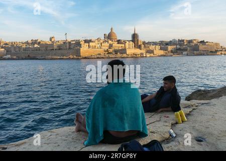 Plage rocheuse de Sliema, deux personnes se détendant loin de la ville de la Valette au coucher du soleil, Malte Banque D'Images