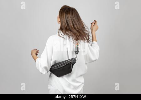 Femme blanche portant un Jean blanc, une chemise et un sac de ceinture en cuir noir croisé sur fond gris Banque D'Images