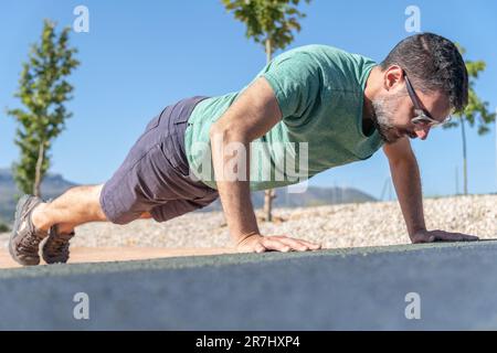 homme aux cheveux sombres vu en profil avec une barbe et des lunettes de soleil faisant des push-up sur un sol en pente Banque D'Images