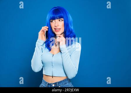 Modèle jeune femme plein de confiance avec des cheveux colorés dans des vêtements décontractés souriant et regardant loin tout en parlant sur un smartphone sur fond bleu Banque D'Images