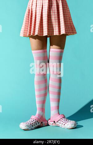 Anonyme jeune femme portant une robe à carreaux et de longues chaussettes à rayures roses avec des chaussures tout en se tenant sur un sol turquoise avec de l'ombre Banque D'Images