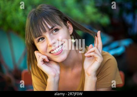 Portrait d'une jeune femme positive avec un sourire ondoyant, en regardant loin avec la main au menton et en touchant les cheveux tout en étant assise sur une chaise contre un arbre vert flou Banque D'Images