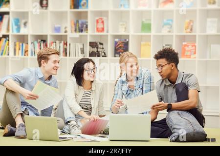 Groupe de contenu confiance jeunes étudiants multiethniques dans des tenues décontractées assis sur le sol dans la bibliothèque et discuter de la tâche ensemble Banque D'Images