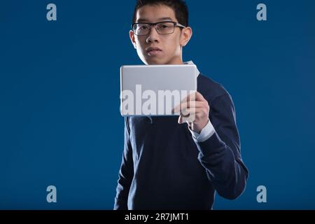 Un homme asiatique regarde les nouvelles sur sa tablette, s'arrêtant pour regarder ici, sur fond bleu. En portant des lunettes, il s'intéresse à ce que l'écran affiche - Banque D'Images