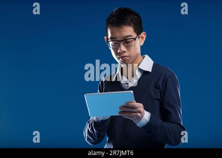 L'homme asiatique, à lunettes, absorbé dans sa tablette, vérifie les nouvelles sur fond bleu. Son intérêt réside dans le contenu de l'écran - vidéo, nouvelles, un e-mail, Banque D'Images