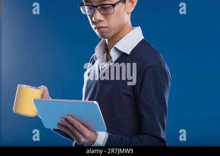 Un homme asiatique consulte les informations sur sa tablette, avec une tasse jaune, sur fond bleu. Il est engrosé dans le contenu de l'écran, que ce soit une vidéo, des nouvelles, Banque D'Images