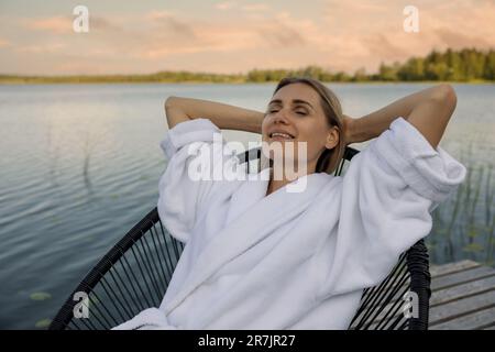 femme en peignoir blanc se détendant sur une chaise sur la passerelle du lac lors d'une chaude soirée d'été. plaisir de la nature Banque D'Images