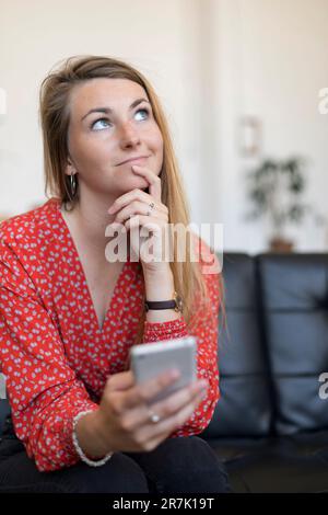 Jeune femme blonde assise sur un canapé avec un téléphone portable dans sa main et regardant avec attention. Banque D'Images