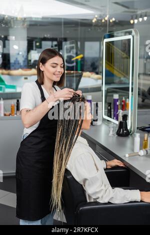 l'industrie de la beauté, les tresses, coiffeur heureux tressant cheveux de femme en salon, processus de tressage, client de salon, profession de beauté, satisfaction de la clientèle, Banque D'Images