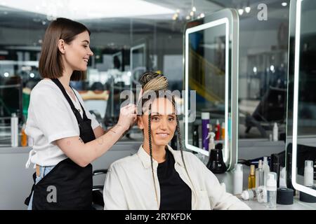 salon de coiffure, coiffeur heureux de faire un pain de cheveux à la cliente féminine avec des braïdes, des femmes gaies, la satisfaction de la clientèle, la cliente dans le salon, salon de beauté servi Banque D'Images