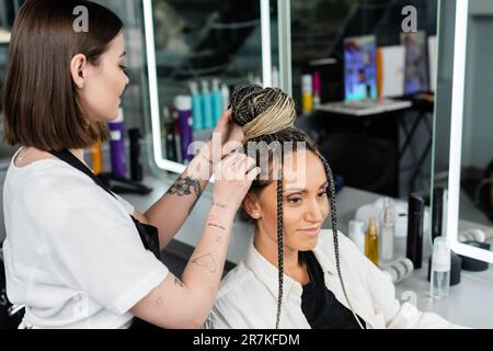 salon de coiffure, coiffeur tatoué faisant un pain de cheveux à la cliente féminine avec des braïdes, des femmes heureuses, la satisfaction de la clientèle, la cliente dans le salon de beauté servic Banque D'Images