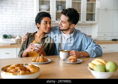 Couple positif amoureux de différentes nationalités, assis et embrassant à la maison dans la cuisine, vêtu de vêtements élégants, boire un café du matin avec des croissants, regarder les uns les autres, sourire Banque D'Images