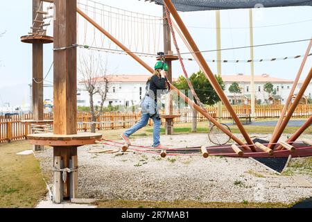 Adolescente dans l'équipement de harnais d'escalade, casque de sécurité vert de sport. Parc d'attractions de corde. Fixation fixant le mousqueton à la corde de sécurité. Hangin Banque D'Images