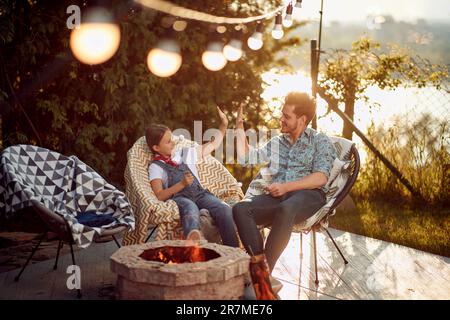 Père et fille de haut fiving, assis à l'extérieur dans leur cour près d'une cheminée en appréciant le temps ensemble, torréfaction des tâches pendant l'été. Style de vie, f Banque D'Images