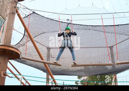 Adolescente dans l'équipement de harnais d'escalade, casque de sécurité vert de sport. Parc d'attractions de corde. Fixation fixant le mousqueton à la corde de sécurité. Hangin Banque D'Images