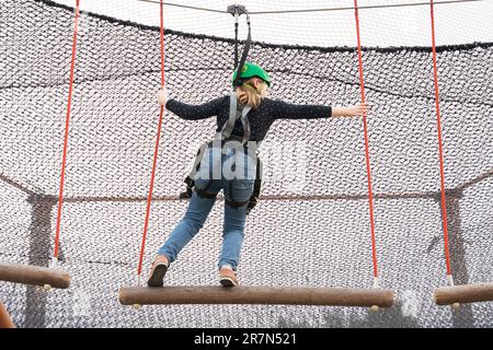 Adolescente dans l'équipement de harnais d'escalade, casque de sécurité vert de sport. Parc d'attractions de corde. Fixation fixant le mousqueton à la corde de sécurité. Hangin Banque D'Images