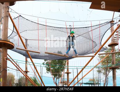 Adolescente dans l'équipement de harnais d'escalade, casque de sécurité vert de sport. Parc d'attractions de corde. Fixation fixant le mousqueton à la corde de sécurité. Hangin Banque D'Images