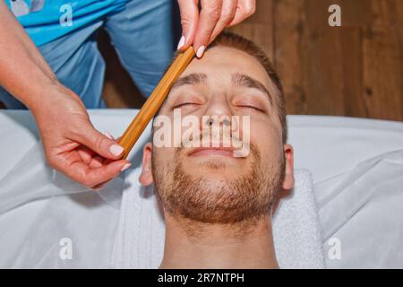 Concept bien-être. Un masseur professionnel offre à un homme un massage du visage thaïlandais avec des bâtons spéciaux dans un centre de spa. Relaxation et récupération après le travail Banque D'Images