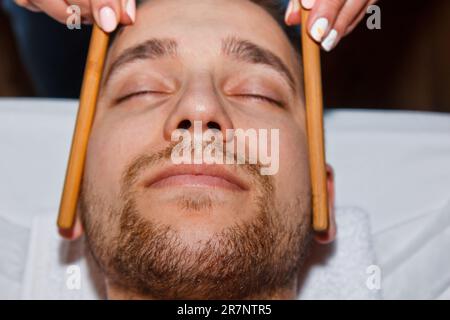 Concept bien-être. Un masseur professionnel offre à un homme un massage du visage thaïlandais avec des bâtons spéciaux dans un centre de spa. Relaxation et récupération après le travail Banque D'Images