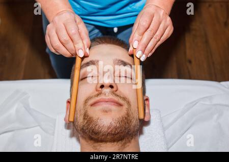 Concept bien-être. Un masseur professionnel offre à un homme un massage du visage thaïlandais avec des bâtons spéciaux dans un centre de spa. Relaxation et récupération après le travail Banque D'Images