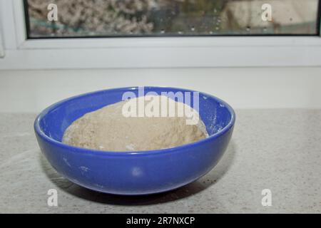 Une pile de pâte à pétrir saupoudrée de farine de blé se trouve dans un bol bleu sur une table de cuisine blanche. cuisson, cuisson. Banque D'Images