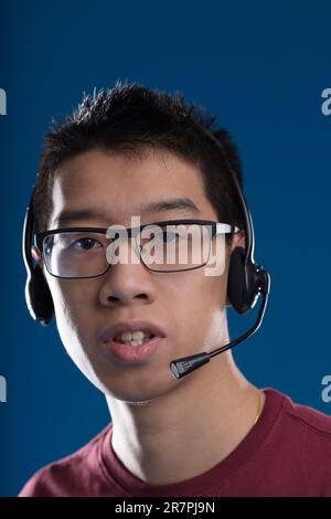 « Comment puis-je vous aider ? » dit un jeune homme avec des lunettes, un casque, travaillant au service à la clientèle. Un opérateur téléphonique ou un joueur sur fond bleu Banque D'Images