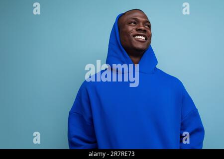 joyeux homme africain de 25 ans dans un élégant sweat à capuche avec capuche sur fond bleu Banque D'Images