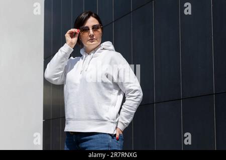 Portrait la femme mûre Brunette en lunettes de soleil porte un sweat à capuche blanc posé sur le mur d'arrière-plan en granit foncé. Sportive confient 40 ans de beau blanc Banque D'Images