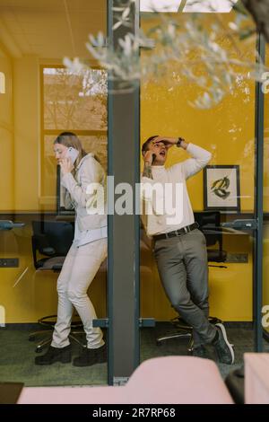 Deux collègues masculins et féminins attrayants ayant un appel téléphonique dans les cabines téléphoniques au bureau Banque D'Images