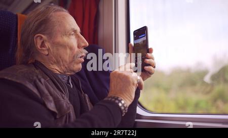 Homme âgé voyageant en train et prenant des photos du paysage par la fenêtre à l'aide d'un smartphone Banque D'Images