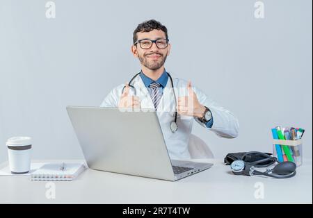Un médecin souriant assis avec un ordinateur portable qui donne les pouces. Un médecin de sexe masculin avec un ordinateur portable montrant les pouces vers le haut en geste ok Banque D'Images